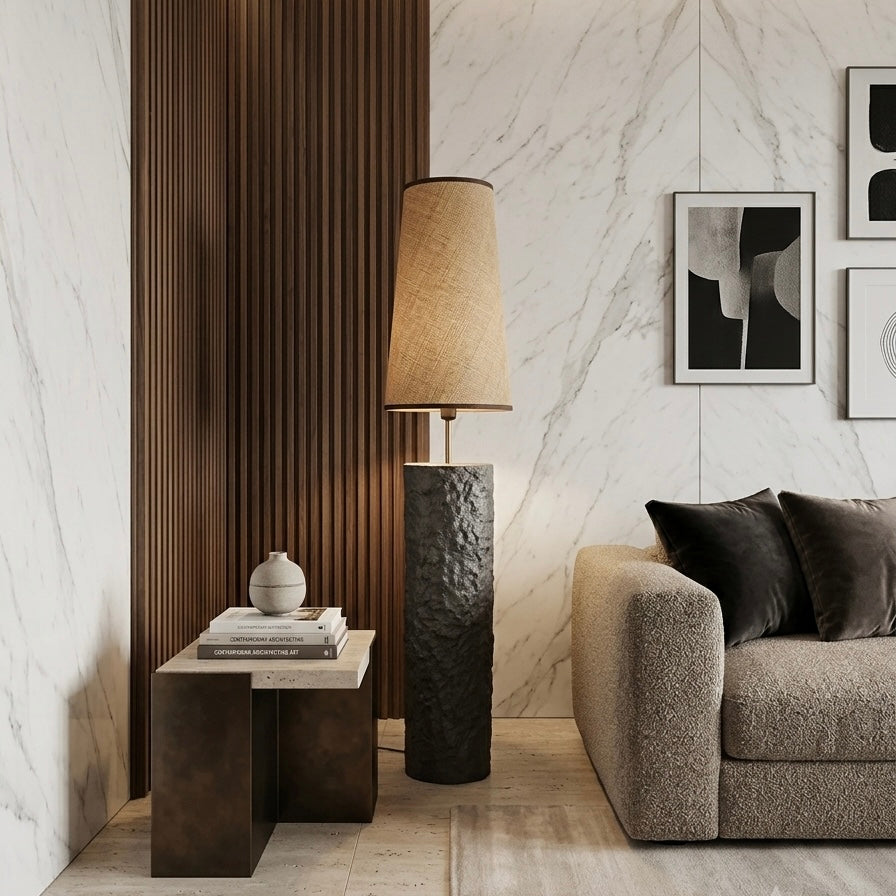 A modern living room corner featuring a textured dark gray cylindrical floor lamp with a beige drum shade, next to a dark wood slatted accent wall and a beige textured sofa.